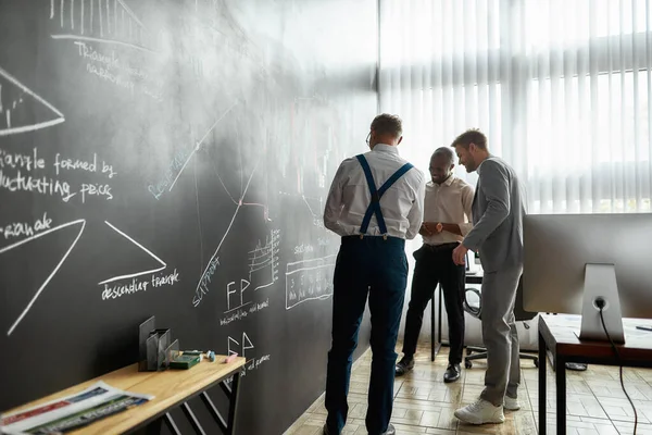 One solution for business education. Full-length shot of diverse employees, traders standing near blackboard full of charts, while discussing the strategy of work. Horizontal shot — Stock fotografie
