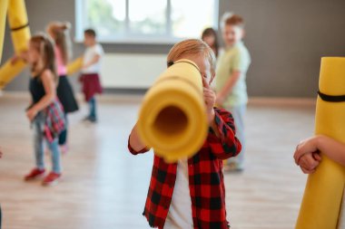 Yoga time. Little boy in checkered shirt holding a yellow mat and looking through the hole. Group of children in casual clothes holding yoga mats while standing in the dance studio