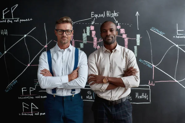 We kennen Financiën beter. Diverse medewerkers, handelaars staan in de buurt van schoolbord vol grafieken en kijken naar de camera, terwijl ze praten over de strategie van het werk. Horizontaal schot — Stockfoto