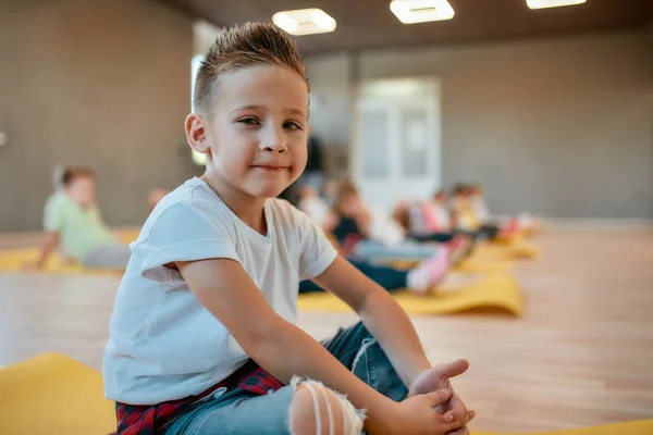 Portrait of a cute little boy in white t-shirt sitting on the yoga mat and smiling at camera while having a yoga class in the dance studio — Stock Photo, Image