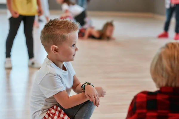一个时髦的小男孩坐在地板上，一边看一边上舞蹈课的画像。舞蹈工作室 — 图库照片