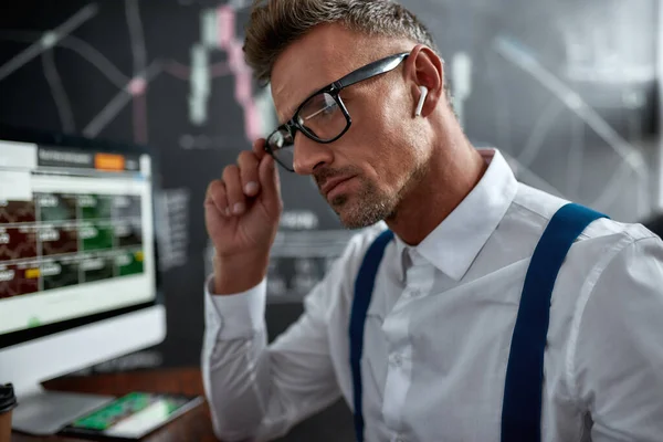 Prenditi cura del tuo futuro. Primo piano di elegante uomo d'affari, commerciante seduto di fronte al monitor del computer mentre si lavora con auricolari wireless. Lavagna ricca di grafici e analisi dei dati in background . — Foto Stock