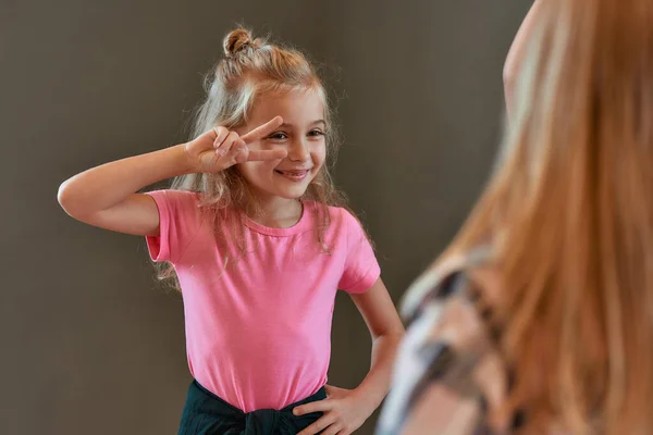 Porträtt av söt liten flicka i rosa t-shirt visar en fredsgest och leende när du står mot grå vägg och har koreografi klass i dansstudion. — Stockfoto