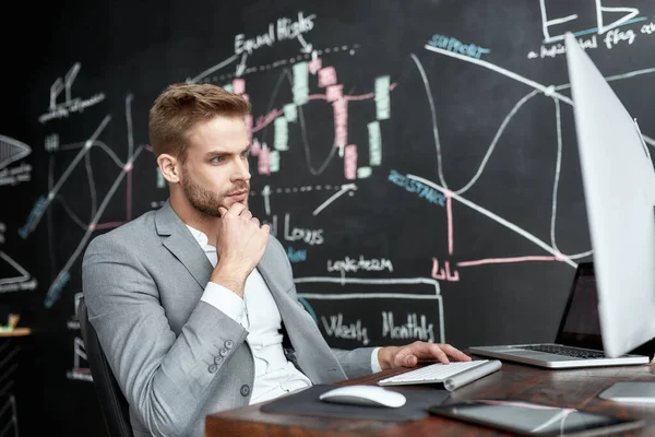 For the real difference. Young trader sitting by desk in front of computer monitor while working in the office. Blackboard full of charts and data analyses in background. — Stock Photo, Image