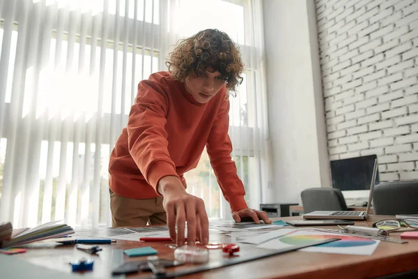 Jonge ontwerper werkt aan een nieuw project. Geconcentreerde jongeman die iets van de tafel nam met veel creatieve dingen erop. Jonge ontwerper in casual kleding werken in het moderne kantoor — Stockfoto