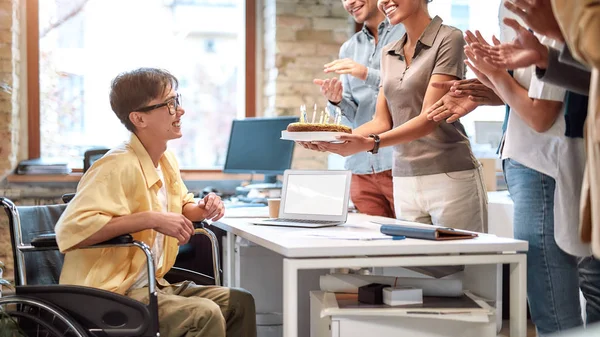 Geburtstagsfeier. Gruppe junger und glücklicher Geschäftsleute gratuliert ihrem Kollegen im Rollstuhl zum Geburtstag im modernen Büro — Stockfoto