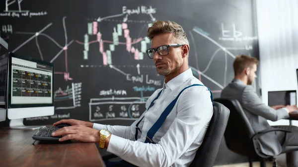 El arte de la negociación. Comerciante de mediana edad sentado junto al escritorio frente al monitor de la computadora mientras trabaja en la oficina. Su colega y pizarra llena de gráficos y análisis de datos en segundo plano . — Foto de Stock