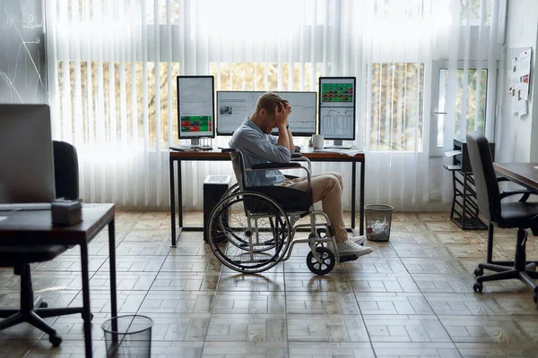 Oh no. Portrait of young upset male trader in a wheelchair holding his head in hands and feeling frustrated while sitting at his workplace in the office