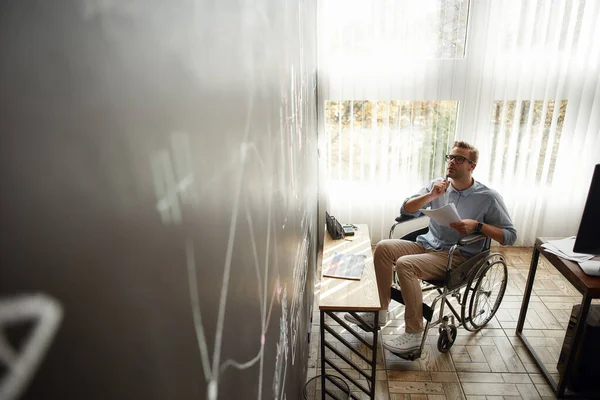 Denkproces. Bedachtzame jonge mannelijke handelaar in een rolstoel in het bezit van een aantal documenten en analyseren op grafieken en grafieken op schoolbord tijdens het werken in het moderne kantoor — Stockfoto