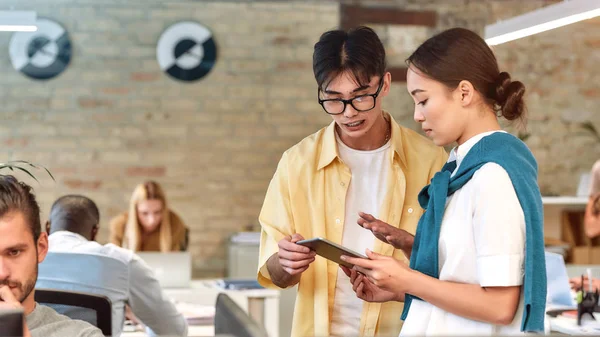 A trabalhar num novo projecto. Dois colegas asiáticos em desgaste casual olhando para tablet digital e discutindo algo enquanto estão juntos no escritório moderno — Fotografia de Stock