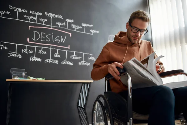 Looking for creative ideas. Portrait of concentrated young designer in a wheelchair reading book while sitting near blackboard with presentation in the creative office