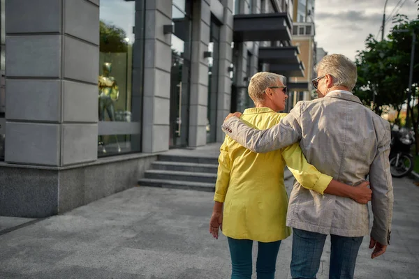 Visão traseira do elegante casal de meia-idade em jaquetas de terno estão abraçando enquanto caminham juntos pela rua da cidade — Fotografia de Stock