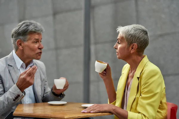 コーヒーの時間だ。モダンな大人のカップルはコーヒーを飲みながら、屋外のカフェに座っている間に話し合います — ストック写真