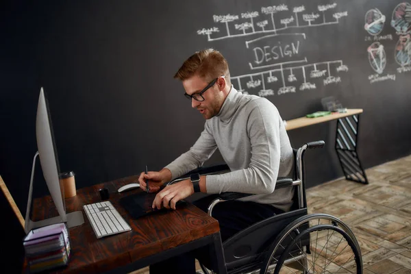 İşiyle meşgul bir tasarımcı. Tekerlekli sandalyedeki genç ve yakışıklı erkek ofis çalışanının tasarım stüdyosunda çalışırken stilus kalem ve dijital tablet kullanışının yan görüntüsü. — Stok fotoğraf