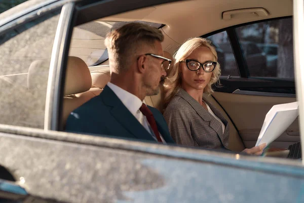 Planificación de un nuevo proyecto. Mujer joven y hermosa en gafas con documentos y escuchando a su socio de negocios mientras está sentada en el coche — Foto de Stock