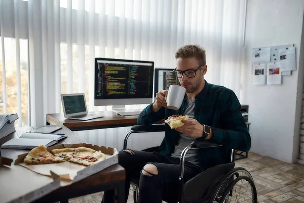 Czuję głód. Młody deweloper sieci w wózku inwalidzkim jedzący pizzę i pijący herbatę siedząc w swoim miejscu pracy w nowoczesnym biurze. Wiele ekranów komputerowych z kodem programu — Zdjęcie stockowe