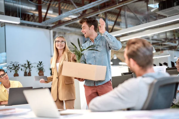 Welkom in ons team. Jonge knappe man in casual wear holding box met dingen en zwaaien naar zijn nieuwe collega 's terwijl ze in het moderne kantoor — Stockfoto