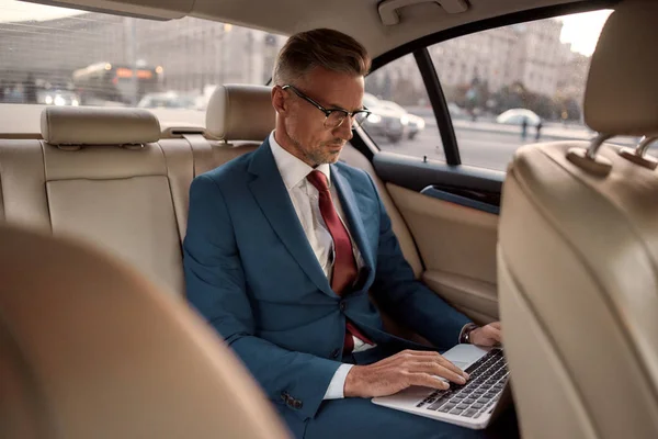 Effectieve bedrijfsstrategie. Zelfverzekerde volwassen zakenman in volle pak werken op zijn laptop, terwijl zitten in de auto — Stockfoto