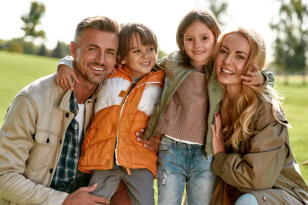 Relajándonos juntos. Retrato de feliz hermosa familia con los niños abrazando y mirando a la cámara con sonrisa mientras pasa tiempo al aire libre —  Fotos de Stock