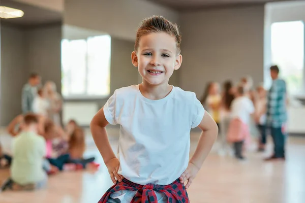 Cool dancer. Portrait of a cute and happy little boy in white t-shirt keeping arms on hips and looking at camera with smile while standing in the dance studio