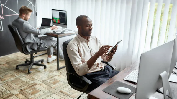 Balancing the best interest. Cheerful african male trader sitting by desk and studying analytical reports using tablet pc in the office. His colleague is working in the background — Zdjęcie stockowe