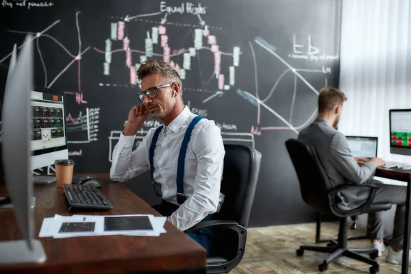 Investi oggi, guadagna domani. Elegante uomo d'affari, commerciante seduto di fronte al monitor del computer mentre si lavora con auricolari wireless. Il suo collega e lavagna piena di analisi dei dati in background . — Foto Stock