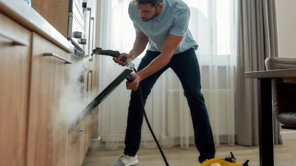 Servizio al lavoro. Giovane uomo in uniforme pulizia forno elettrico con pulitore a vapore mentre lavora nella cucina moderna — Foto Stock