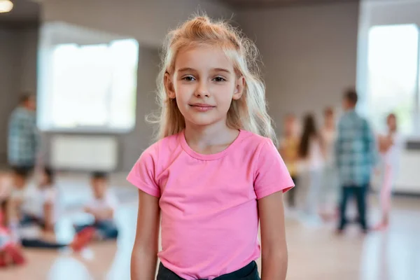 Ritratto di una graziosa bambina in t-shirt rosa che guarda la macchina fotografica mentre è in piedi nello studio di danza — Foto Stock