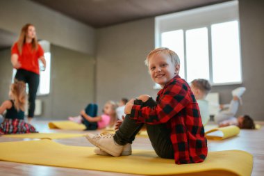 Positive little boy in casual clothes sitting on the mat and smiling at camera. Group of children doing exercises with female trainer in the dance studio
