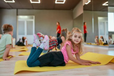 Çocuklar ve spor. Dans stüdyosunda yoga minderinde yatarken kameraya gülümseyen mutlu küçük bir kızın yan görüntüsü. Bir grup çocuk, genç bayan eğitmenle esneme egzersizleri yapıyor.