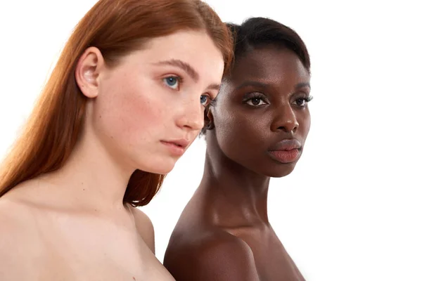 Černobílý. Boční pohled na dvě multikulturní mladé ženy se zářící pletí, které se dívají jinam, zatímco pózují ve studiu nad bílým pozadím. Krásné modely s jinou barvou pleti stojící vedle sebe — Stock fotografie