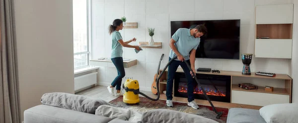 Wij regelen de puinhoop. Twee professionele schoonmakers in uniform werken in de woonkamer. Jonge blanke man maakt het tapijt schoon met stofzuiger, afro-Amerikaanse vrouw veegt stof van de planken — Stockfoto