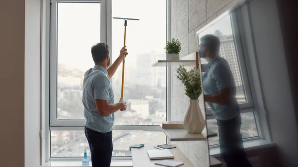 Rápido y eficiente. Limpiador masculino en ventana de limpieza uniforme con escobilla mientras trabaja en la sala de estar —  Fotos de Stock