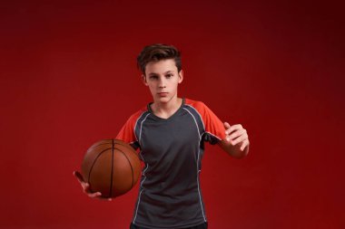 İnan ve Başar. Genç bir çocuk sporla meşgul, basketbol tutarken kameraya bakıyor. Kırmızı arka planda izole edilmiş. Fitness, eğitim, aktif yaşam tarzı konsepti. Yatay çekim.