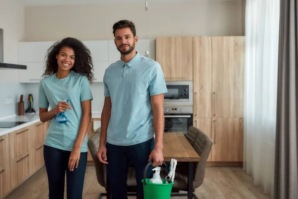 Profesjonalny zespół sprzątaczy. Młody biały mężczyzna i afro amerykańska kobieta trzymając detergenty, patrząc w obiektyw i uśmiechając się podczas sprzątania w kuchni. Profesjonalne środki czystości w mundurze — Zdjęcie stockowe