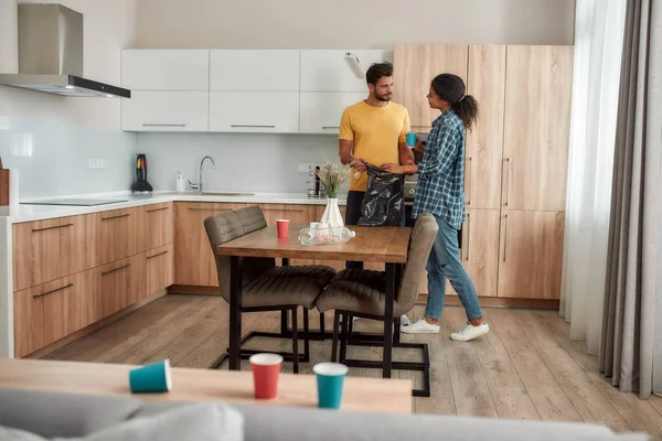 Már majdnem kész. Fiatal multikulturális pár alkalmi ruhában, szemeteszsákot tartva, miközben együtt takarítják a modern konyhát. — Stock Fotó