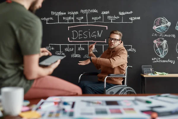 Condividere idee interessanti. Giovane designer UX UI maschio su una sedia a rotelle discutendo qualcosa con il suo collega mentre seduto vicino alla lavagna con presentazione in ufficio moderno — Foto Stock