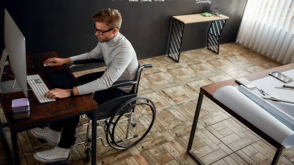 Drukke werkdag. Jonge en creatieve mannelijke kantoormedewerker in een rolstoel met behulp van stylus pen en digitale tablet tijdens het werken op zijn werkplek in het moderne kantoor — Stockfoto