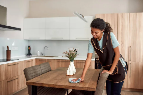 Pur et propre. Jeune femme de ménage afro-américaine positive dans la table de nettoyage de tablier dans la cuisine moderne. Nettoyeur professionnel féminin au travail — Photo