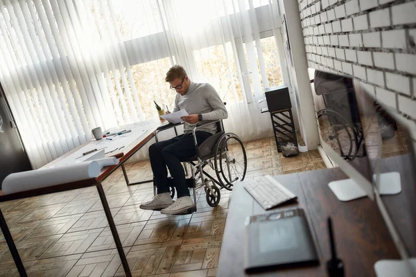 Portret van een jonge mannelijke kantoormedewerker in een rolstoel die iets tekent terwijl hij op zijn werkplek zit in het moderne heldere kantoor — Stockfoto
