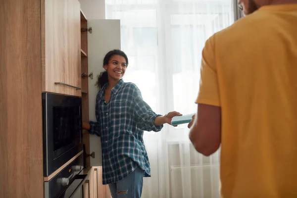 Rób swoje. Wesoła afro amerykańska kobieta w ubraniach casual dając szmatę do swojego chłopaka lub męża podczas sprzątania ich nowoczesnej kuchni razem. Sprzątanie domu — Zdjęcie stockowe