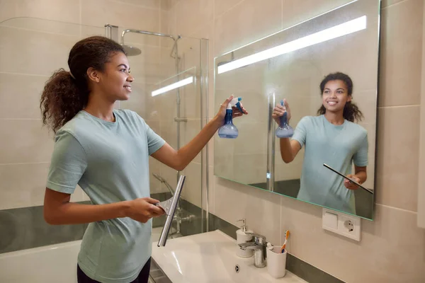 Fazendo a sua casa brilhante limpo. Jovem afro-americana sorrindo em uniforme limpando um espelho com espremedor e detergente spray enquanto trabalhava no banheiro — Fotografia de Stock