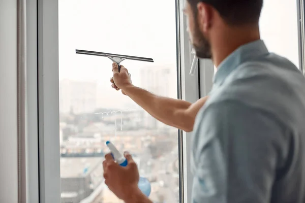 Experto en trabajo. Joven hombre caucásico en ventana de limpieza uniforme con escobilla y detergente en aerosol mientras trabaja en la sala de estar —  Fotos de Stock