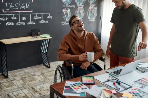 Geweldige stemming voor het werk. Jonge en gelukkige mannelijke Ux Ui ontwerper in een rolstoel met een kop koffie en lachen tijdens het bespreken van een aantal ideeën met zijn collega in het creatieve kantoor — Stockfoto