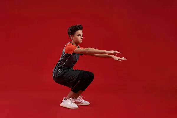 Dedication. A teenage boy is engaged in fitness, he is looking aside while doing sit-ups. Isolated on red background. Sport, training, fitness, active lifestyle concept. Horizontal shot — Stock Photo, Image