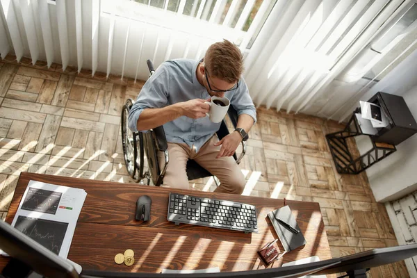 Taking a break. Top view of young businessman or trader in a wheelchair drinking tea or coffee while sitting at his desk in the bright modern office — Stock Photo, Image