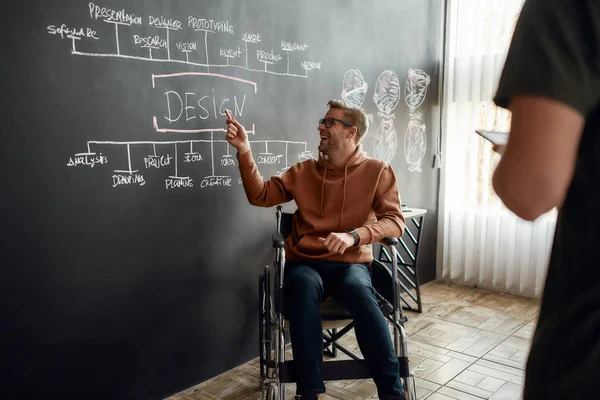 Teamwork. Portret van een jonge positieve ontwerper in een rolstoel die iets bespreekt met zijn collega terwijl hij bij het schoolbord zit met presentatie in het moderne kantoor — Stockfoto