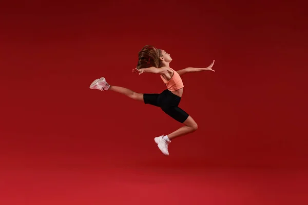 Приложи все усилия. Симпатичный малыш, девочка занимается спортом, она в движении, прыгает в воздухе. Изолированный на красном фоне. Фитнес, тренировки, концепция активного образа жизни. Горизонтальный выстрел — стоковое фото