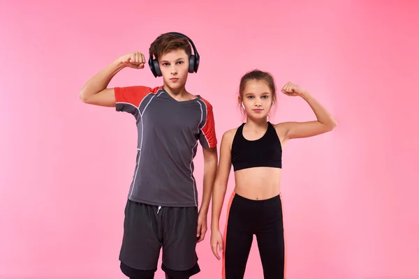 Kita adalah tim. Dua remaja laki-laki dan perempuan mendengarkan musik melalui earphone bersiap-siap untuk berolahraga, menunjukkan bisep terisolasi di latar belakang merah muda. Fitness, olahraga, pelatihan, konsep gaya hidup aktif — Stok Foto