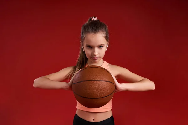 Maradj motivált. Egy aranyos gyerek, sportoló lány, a kosárlabdával való edzés közben a kamerába néz. Piros háttérrel elszigetelve. Fitness, tréning, aktív életmód koncepció. Vízszintes lövés — Stock Fotó